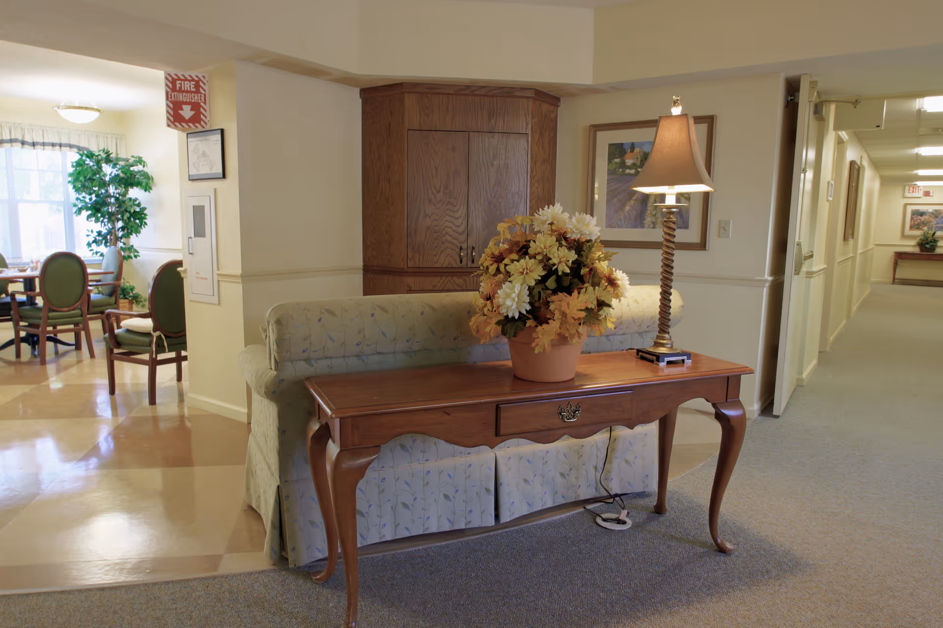 A cozy interior hallway area in a senior living facility featuring a wooden table with a potted flower arrangement and a lamp on it. Behind the table is a cushioned bench with floral upholstery. To the left, there is a dining area with green cushioned chairs and a large window letting in natural light. The hallway extends to the right with framed artwork on the walls and an exit sign visible in the distance.