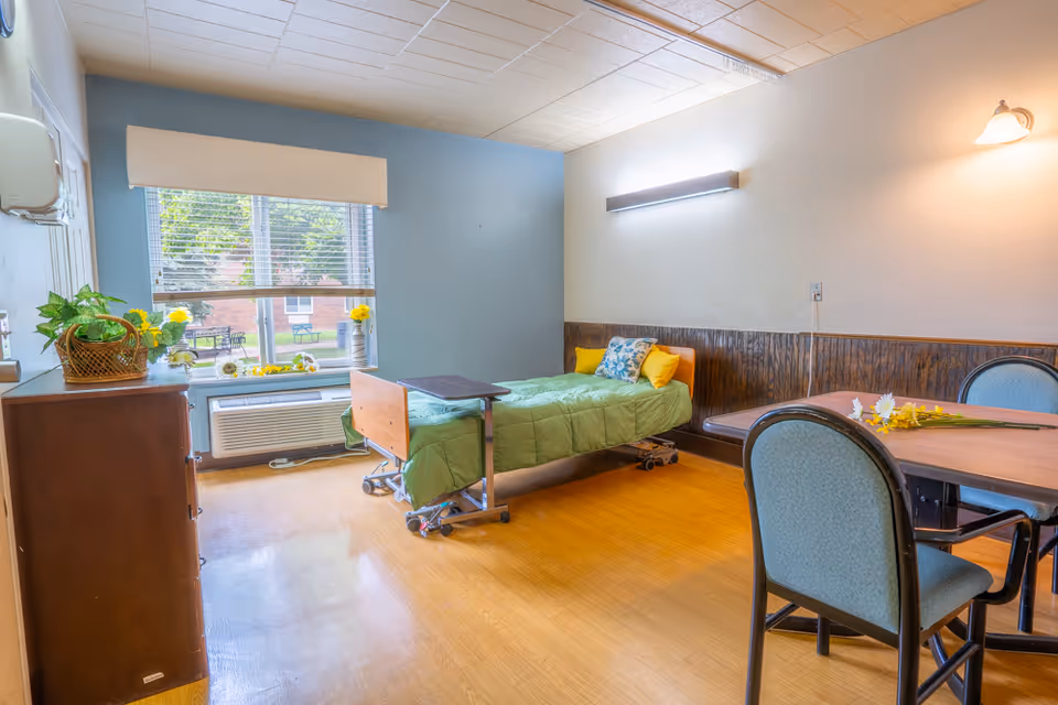 A bright and clean nursing facility room with a single hospital bed covered in a green blanket and yellow pillows. There is a wooden dresser with a basket of plants and flowers on top, a large window with blinds letting in natural light, and a table with chairs and flowers on it. The walls are painted light blue and white with wood paneling along the lower half.