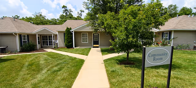 Single-story cottage building with a central sidewalk, grassy lawn, and a 'Community Room' sign in front.
