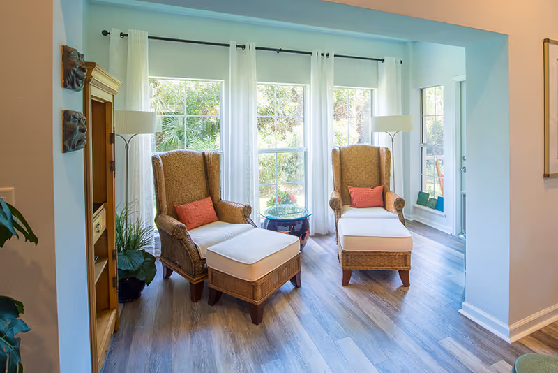 Bright sunroom with two wicker armchairs and matching ottomans facing large windows with sheer curtains.