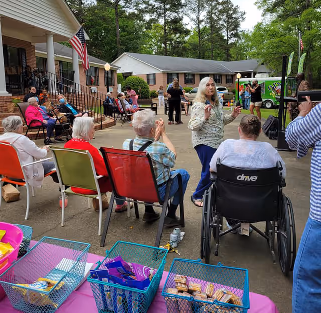 A group of elderly people seated outdoors in chairs and a wheelchair, watching a woman standing and speaking. The setting appears to be a courtyard of an assisted living facility with a building and trees in the background. There are baskets with snacks on a table in the foreground.