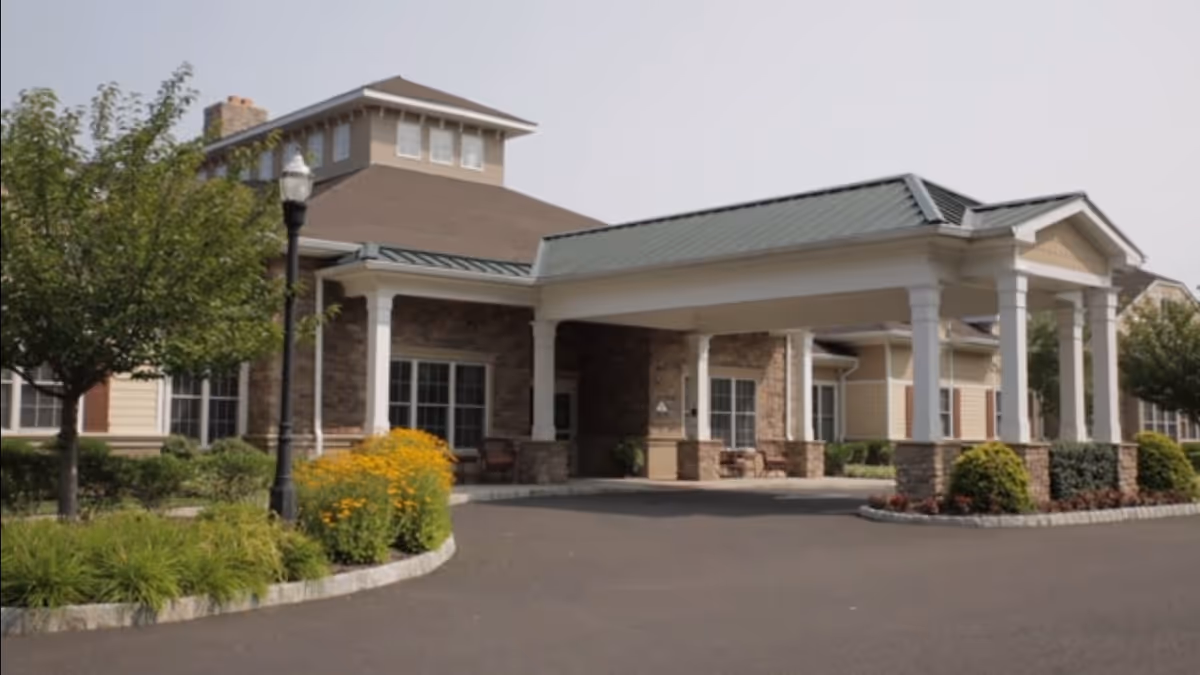 Exterior view of a senior living facility with a covered entrance supported by white columns, stone and beige siding walls, landscaped bushes and flowers, and a paved driveway.
