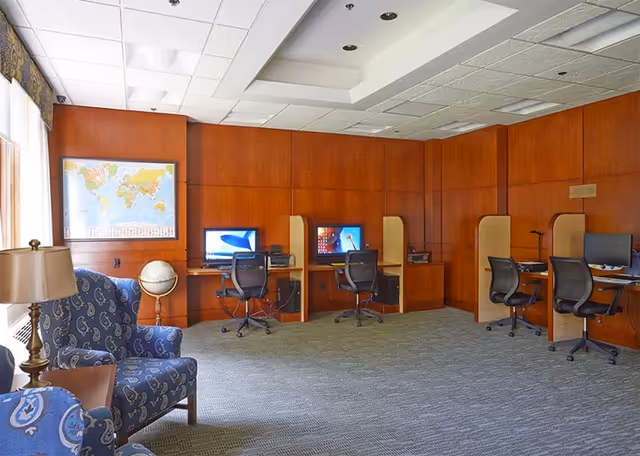 A quiet computer room with four workstations, each equipped with a computer and office chair. The room has wood-paneled walls, a world map on one wall, a globe on a stand, and two blue patterned armchairs with a small table and lamp near a window.