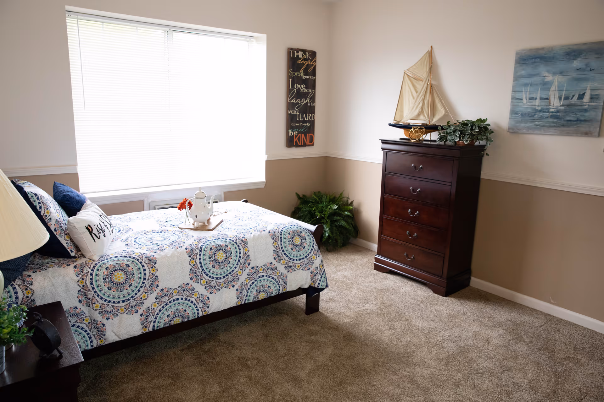 A cozy bedroom with a bed covered in a patterned quilt featuring circular designs in blue, yellow, and white. The bed has several pillows, including one with the word 'Rise' on it. A wooden nightstand with a lamp and a small plant is next to the bed. Across from the bed is a dark wooden chest of drawers topped with a model sailboat and a small plant. The walls are painted white on the upper half and beige on the lower half, with a window covered by white blinds letting in natural light. There is a decorative sign on the wall with inspirational words and a painting of sailboats on the adjacent wall. The floor is carpeted in a light brown color.