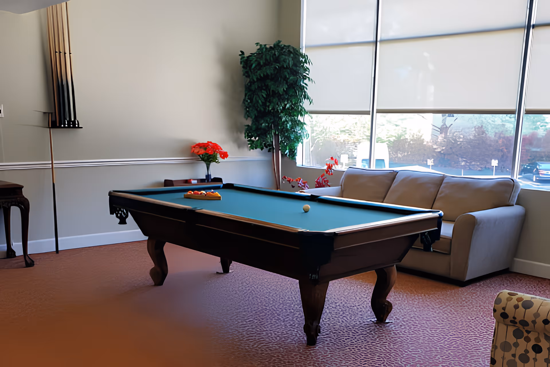 A cozy room with a pool table in the center, a beige sofa by large windows with blinds partially drawn, a tall green plant in the corner, a small table with a vase of red flowers, and a rack holding pool cues on the wall.