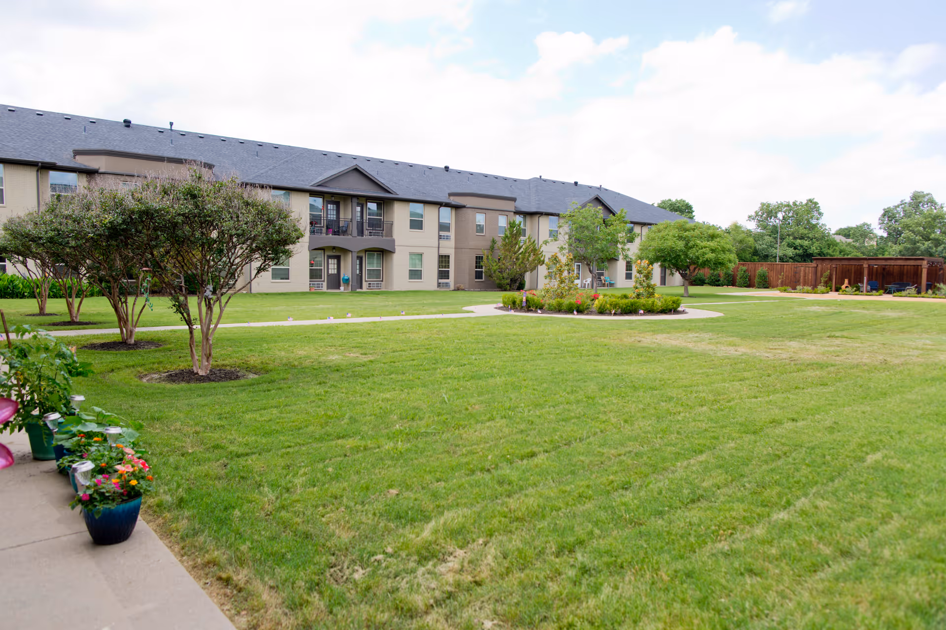 A large, well-maintained grassy courtyard with small trees and flower beds, surrounded by a two-story residential building under a partly cloudy sky.