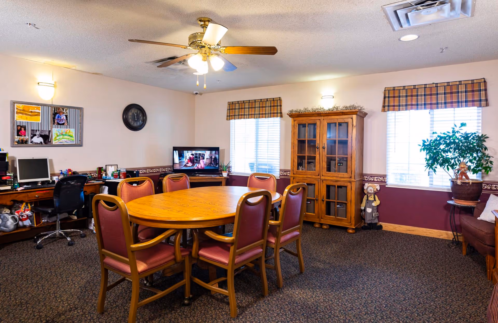 A bright communal dining/activity room with a round wooden table and chairs, a TV, computer desk, and a wooden cabinet.