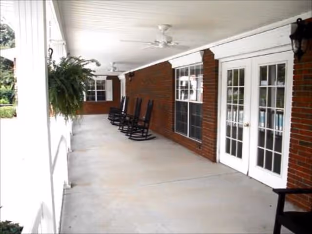 Covered brick porch with several rocking chairs, ceiling fans, hanging plants, and glass double doors.