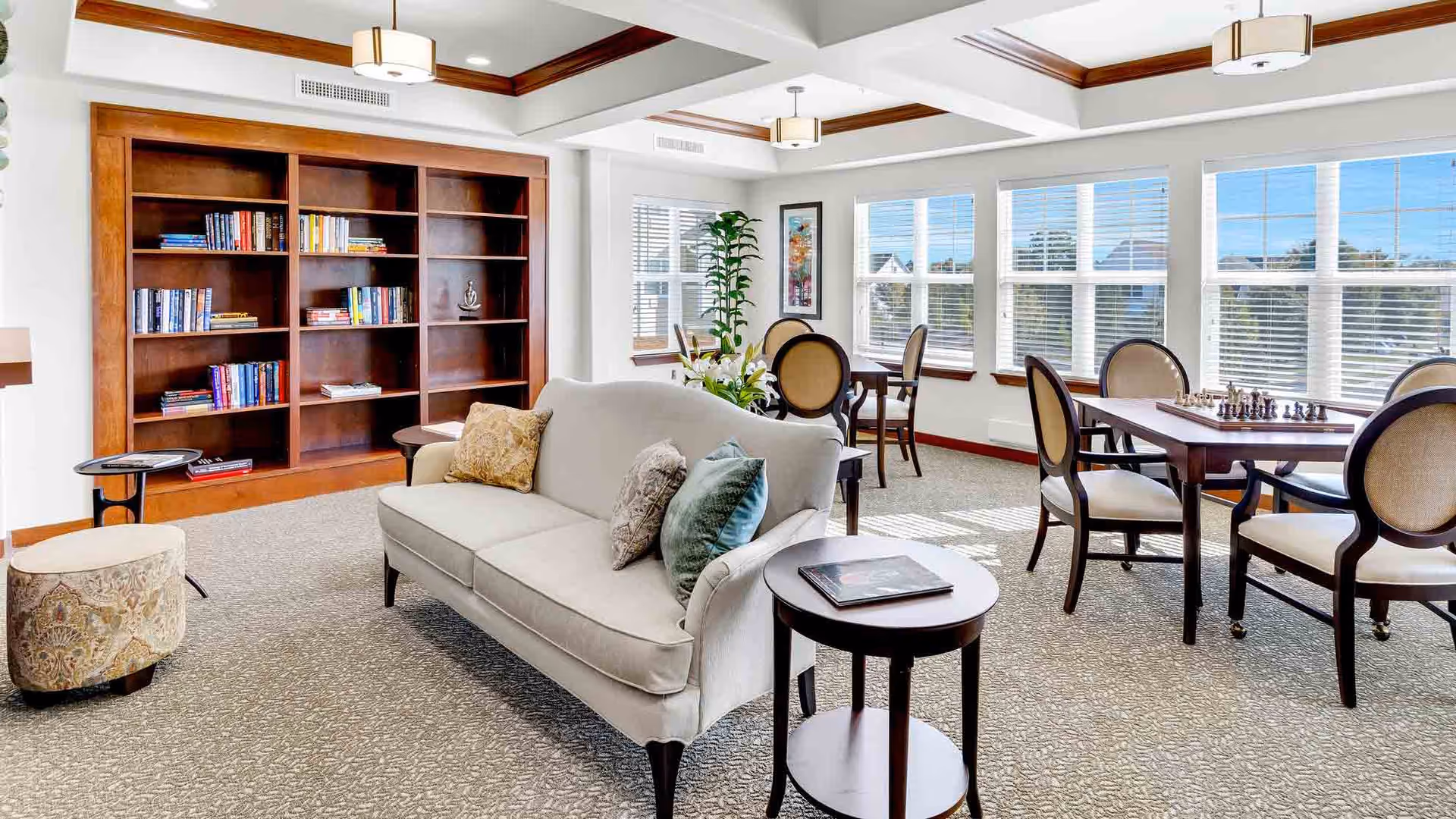 A bright and spacious common area in a senior living facility featuring a beige sofa with decorative pillows, a round side table with a book, a patterned ottoman, a large wooden bookshelf with books, and several tables with chairs near large windows letting in natural light. One table has a chess set on it.