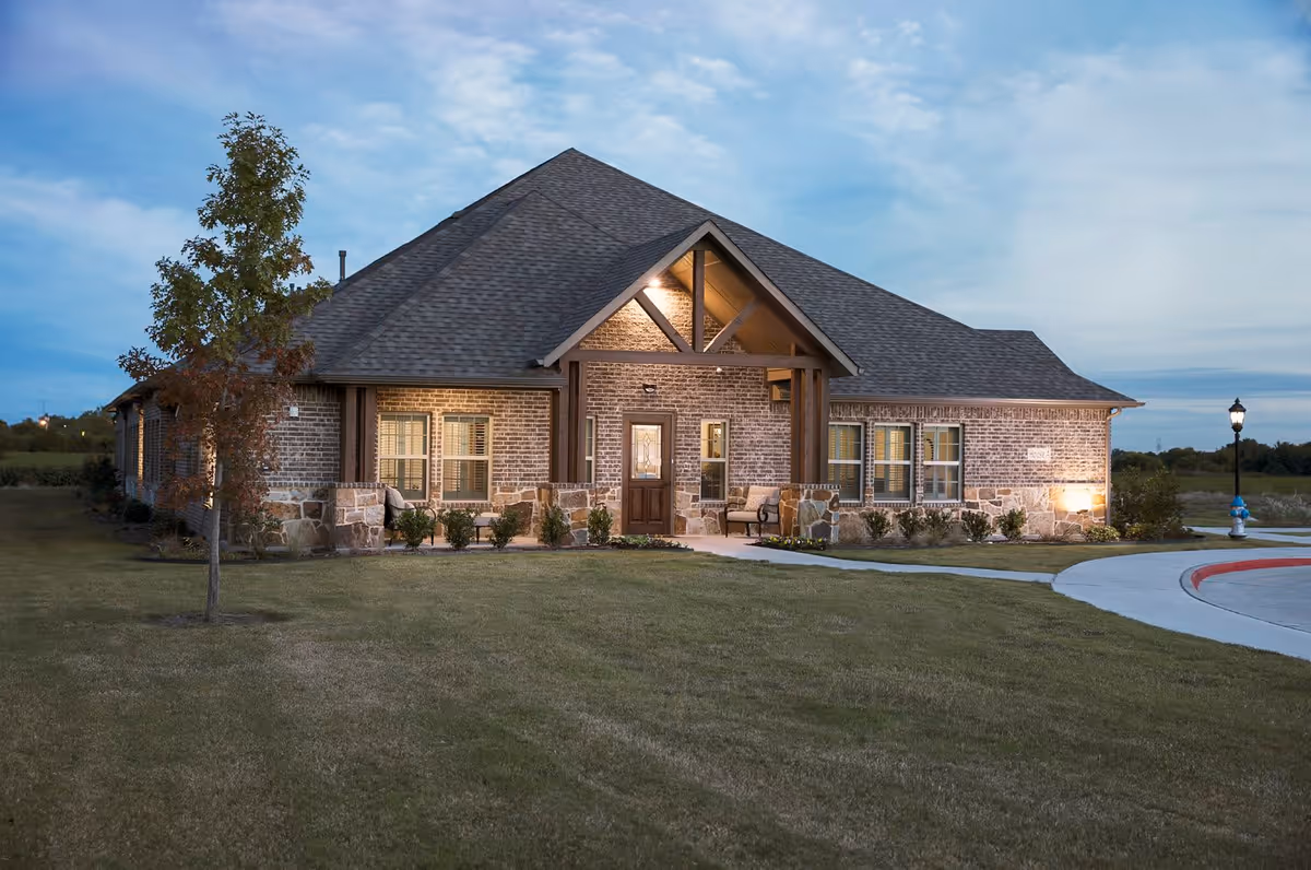 Exterior view of a single-story brick building with a pitched roof at dusk. The building has a covered entrance with wooden beams, several windows with shutters, and a small landscaped area with bushes and a tree in front. A curved driveway and a street lamp are visible on the right side.