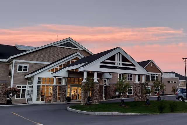 Front entrance of a large senior living facility with a covered porte-cochère, stone columns, and a sunset sky.