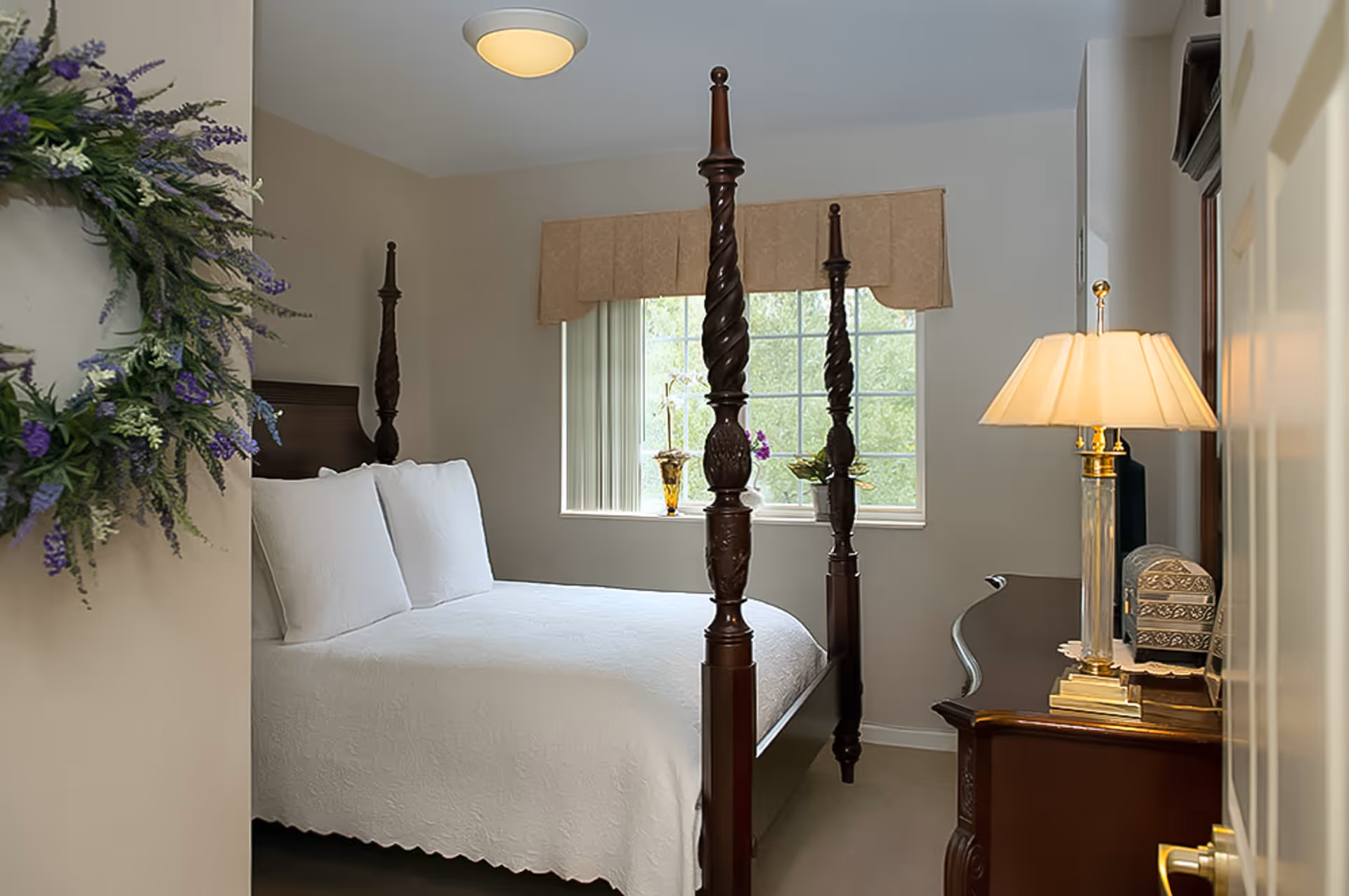A neatly made bed with a white quilt and two white pillows in a bedroom with a four-poster wooden bed frame. There is a window with beige valance letting in natural light, a wooden dresser with a lamp and decorative items, and a floral wreath hanging on the wall near the door.