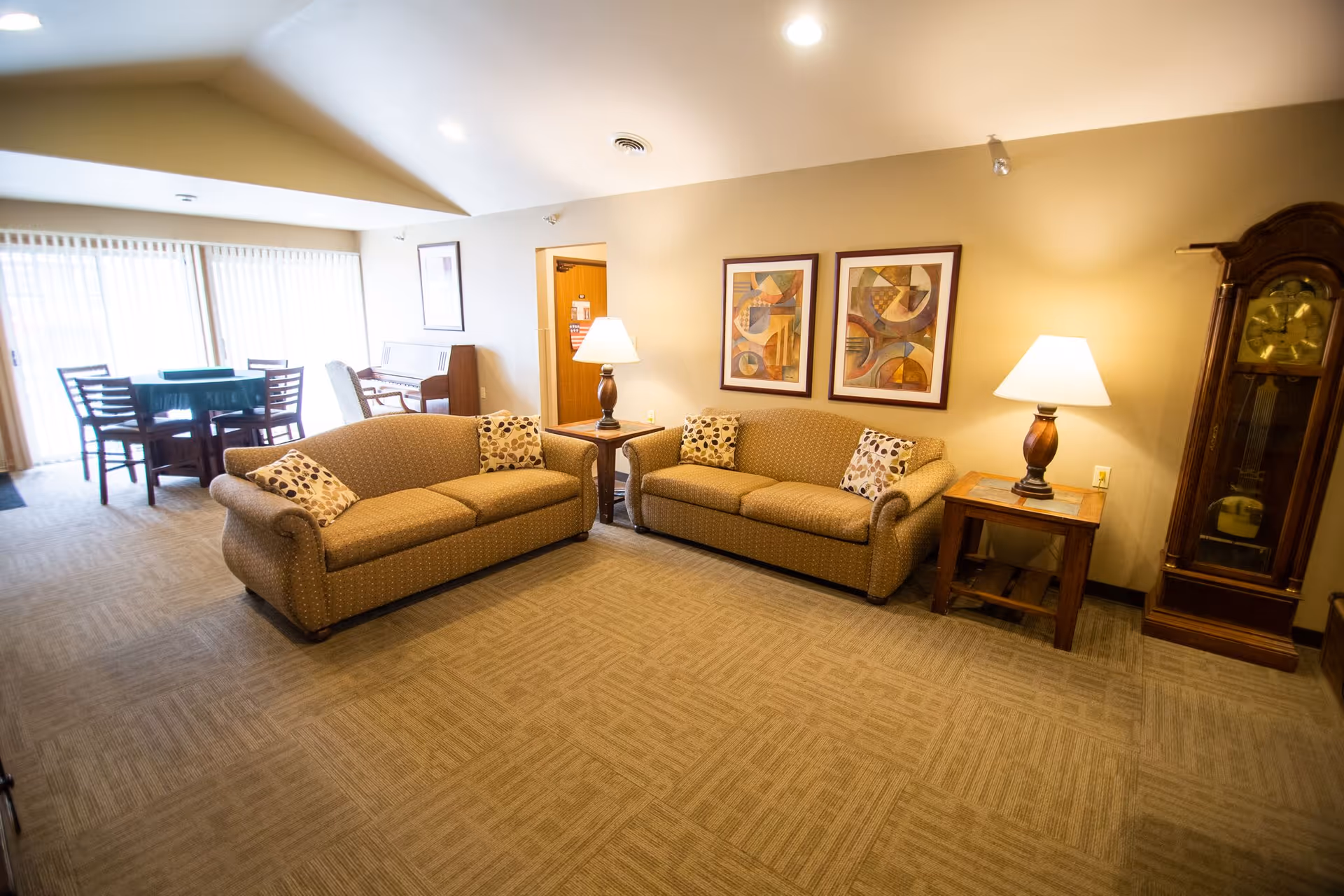 A cozy living room area with two beige patterned sofas facing each other, each with decorative pillows. Between the sofas are wooden side tables with lamps. On the wall above the sofas are two abstract framed paintings. To the right is a tall wooden grandfather clock. In the background, there is a dining table with chairs near large windows covered with vertical blinds, and a piano against the wall.