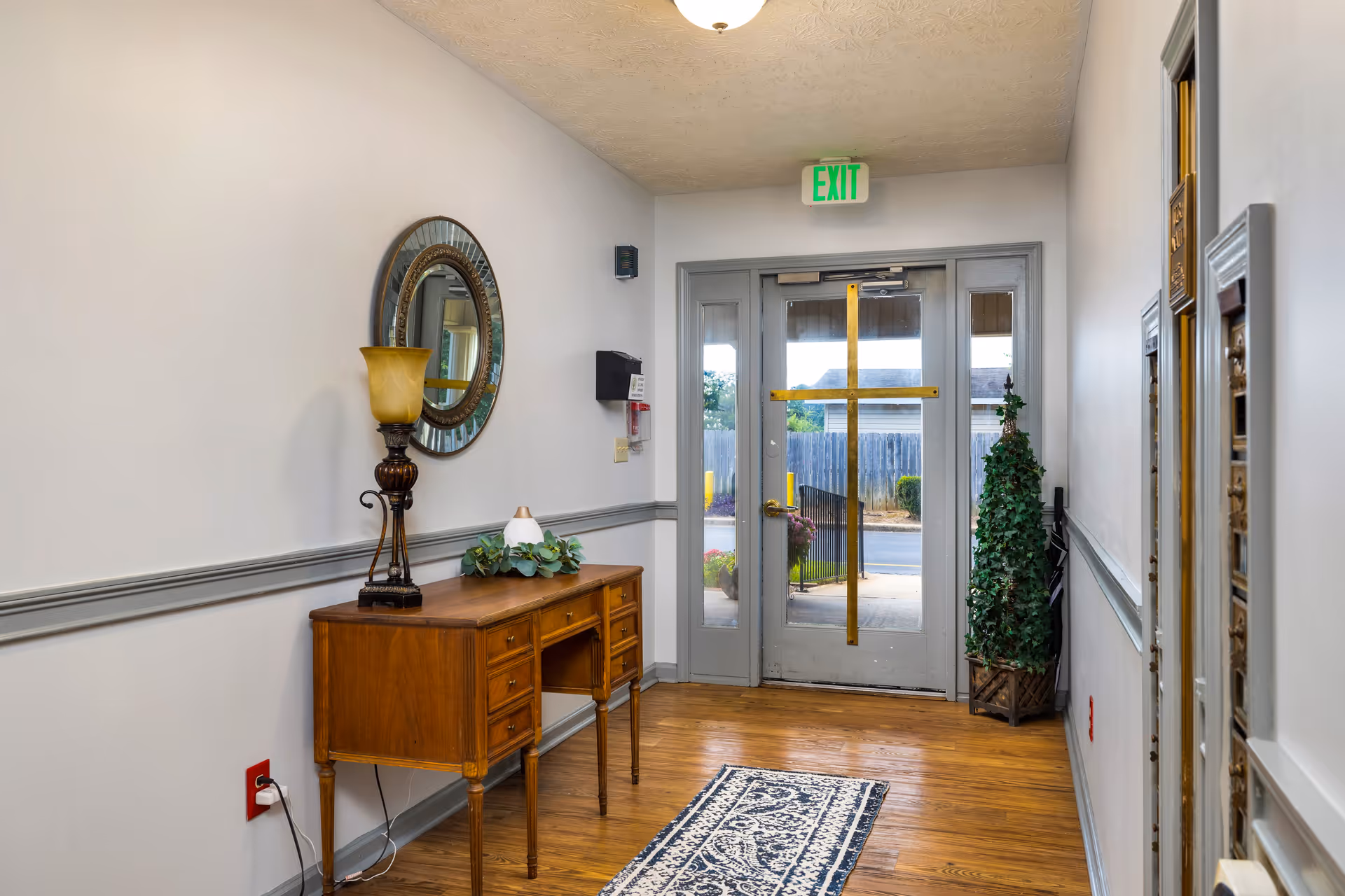 A hallway with wooden flooring and light gray walls featuring a wooden desk with a decorative lamp and a small plant on top. A round mirror hangs above the desk. At the end of the hallway is a glass door with a large gold cross taped on it, an exit sign above, and a potted plant to the right side of the door.