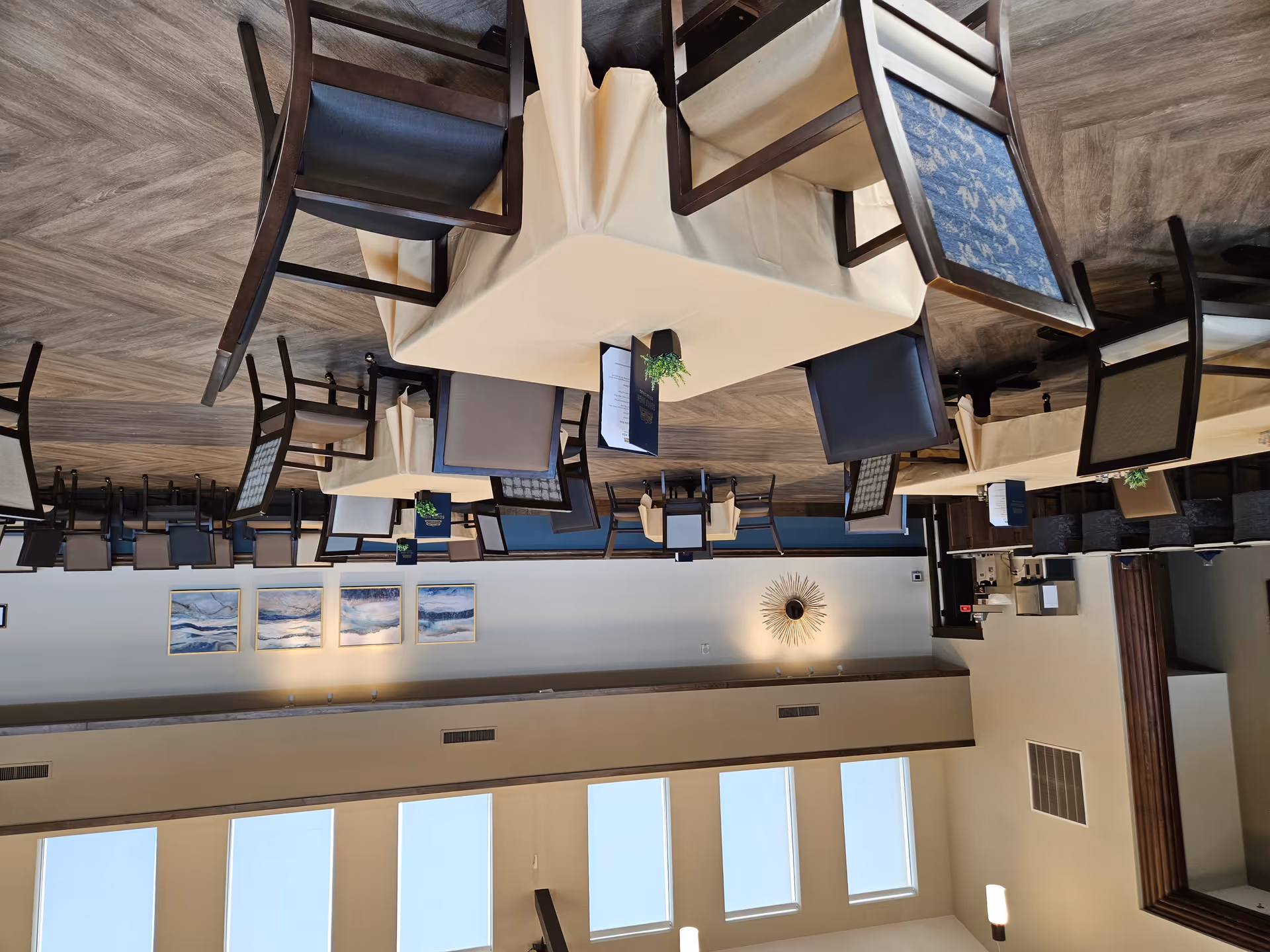 A spacious dining room with multiple tables covered in beige tablecloths, each surrounded by chairs with different upholstery patterns. The room features large windows near the ceiling allowing natural light to fill the space. There are decorative wall art pieces and a sunburst mirror on the walls. A counter with bar stools and a beverage station is visible on the left side.