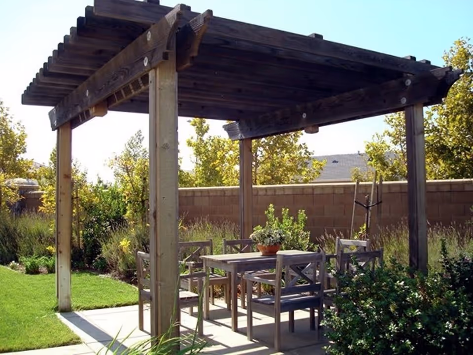 Wooden pergola shading a patio table and chairs in a garden with lawn and shrubs.