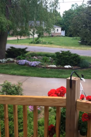 View from a wooden porch railing overlooking a garden with red flowers, a sidewalk, a grassy area with bushes and purple flowers, a road, and trees in the background.