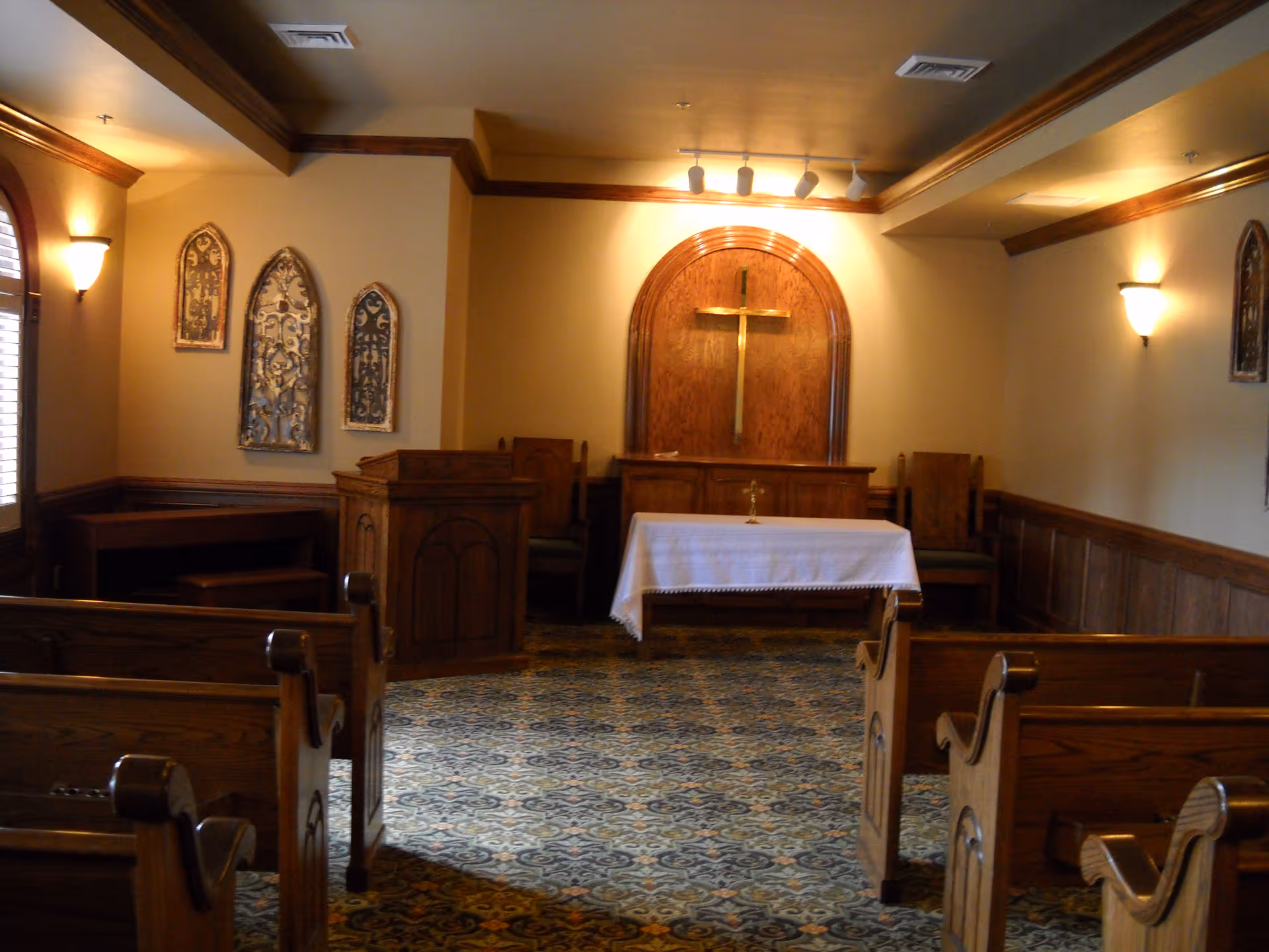 Small chapel interior with wooden pews facing an altar topped by a cross and decorative wall panels.