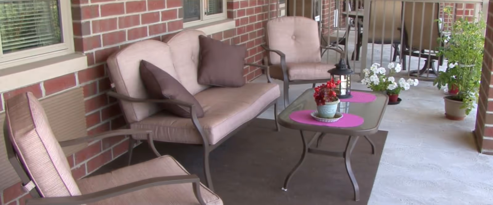 Outdoor patio area with cushioned seating including a loveseat and two chairs around a glass-top coffee table. The table has two pink placemats, a small potted plant, and a lantern. There are several potted plants along the edge of the patio, and the background shows a brick wall with windows.