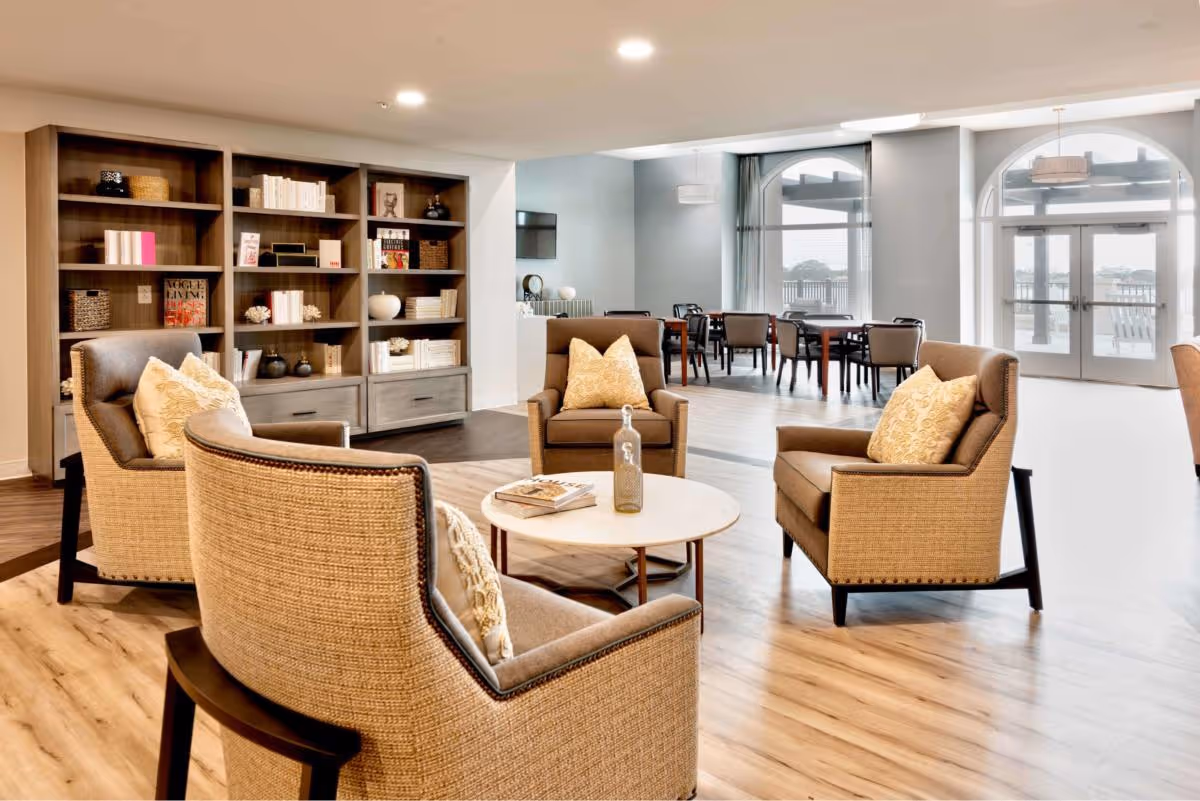 A bright and spacious living area in an assisted living facility featuring four cushioned armchairs arranged around a round coffee table with a decorative bottle and books. Behind the seating area is a large wooden bookshelf filled with books and decorative items. In the background, there are dining tables and chairs near large arched windows and glass doors that let in natural light.