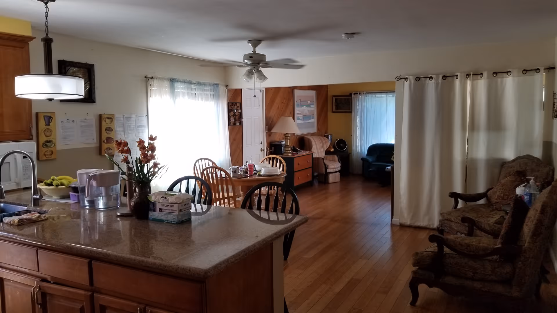 Open-plan interior showing a kitchen island in the foreground, a dining table with chairs in the middle, and a living area with armchairs and curtains in the background.