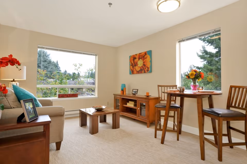 A bright living room with beige walls and carpet, featuring a beige sofa with turquoise pillows, a wooden coffee table, a wooden sideboard with decorative items, and a small round wooden dining table with two chairs. Large windows show green trees outside, and a colorful flower painting hangs on the wall.