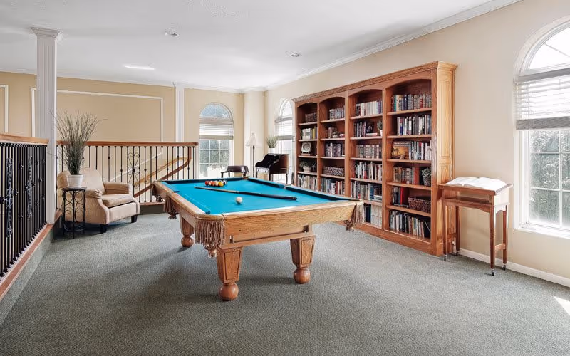 A bright room with a pool table in the center, a large wooden bookshelf filled with books against the wall, a beige armchair with a side table and plant, a piano near the windows, and large arched windows letting in natural light.