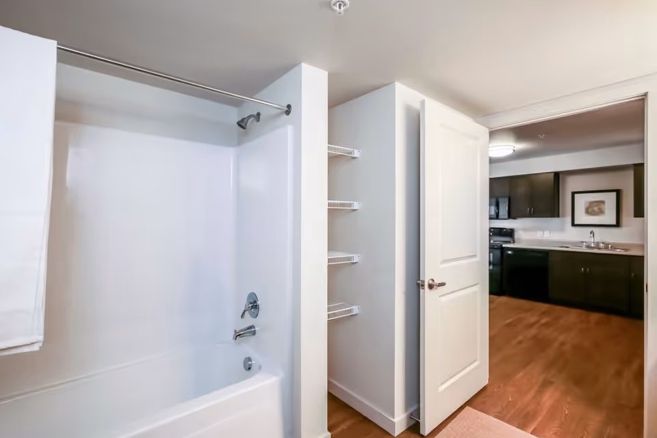 A clean bathroom with a white bathtub and shower combination, a towel hanging on the left side, and an open door leading to a kitchen area with dark cabinets and wooden flooring.