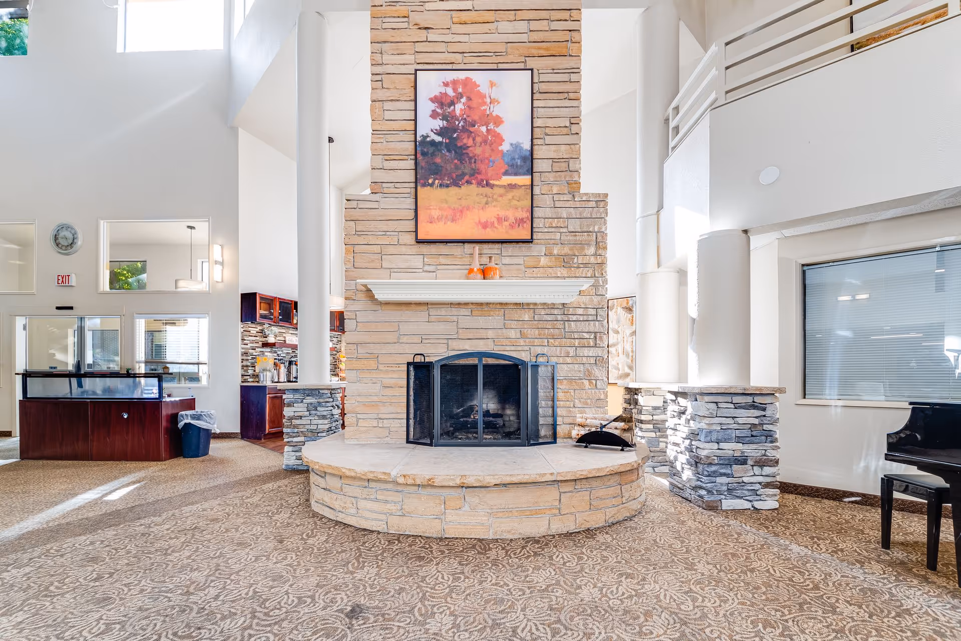 A spacious and bright common area featuring a large stone fireplace with a black metal screen. Above the fireplace hangs a framed painting of red and orange trees in a field. The room has high ceilings, white walls, and patterned carpet flooring. To the left, there is a reception desk and a kitchen area with dark wood cabinets and a tiled backsplash. To the right, a black piano is partially visible near a window with closed blinds. Stone pillars support a balcony railing above.