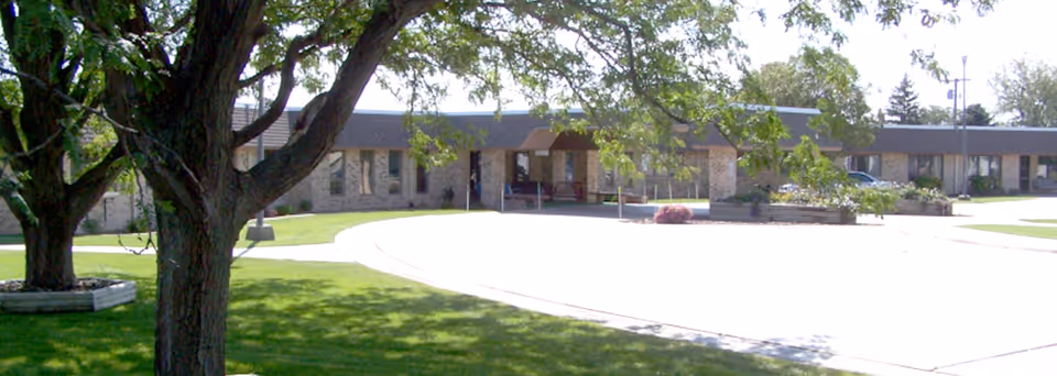 Exterior view of a single-story brick building with a covered entrance, surrounded by green grass and trees on a sunny day.
