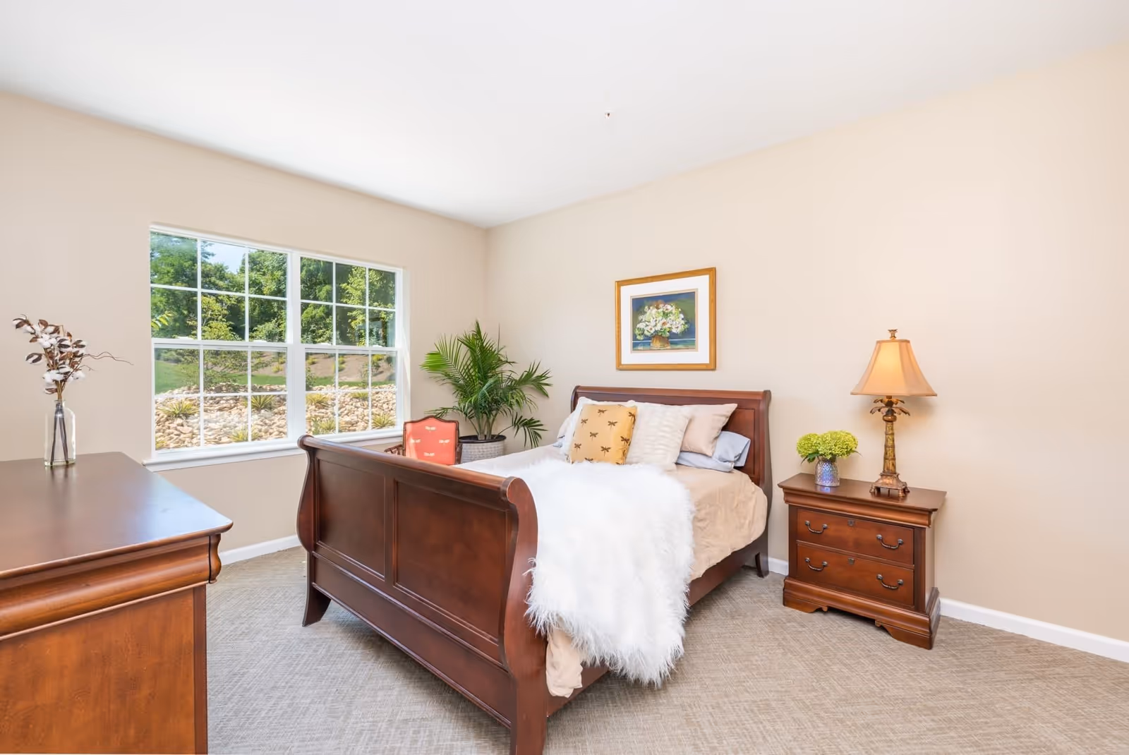 Bright bedroom with a wooden sleigh bed, nightstand and lamp, dresser, a large window showing greenery, and decorative plants.