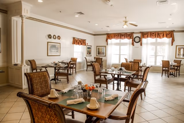 A bright dining room with several wooden tables and chairs arranged neatly. Each table is set with placemats, napkins, cups, glasses, and small flower arrangements. The room has large windows with sheer white curtains and orange valances, framed artwork on the walls, and a ceiling fan with lights.
