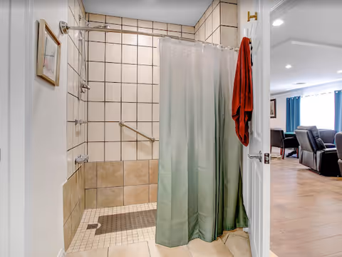 Accessible bathroom with tiled shower and curtain, opening to a living area with seating.