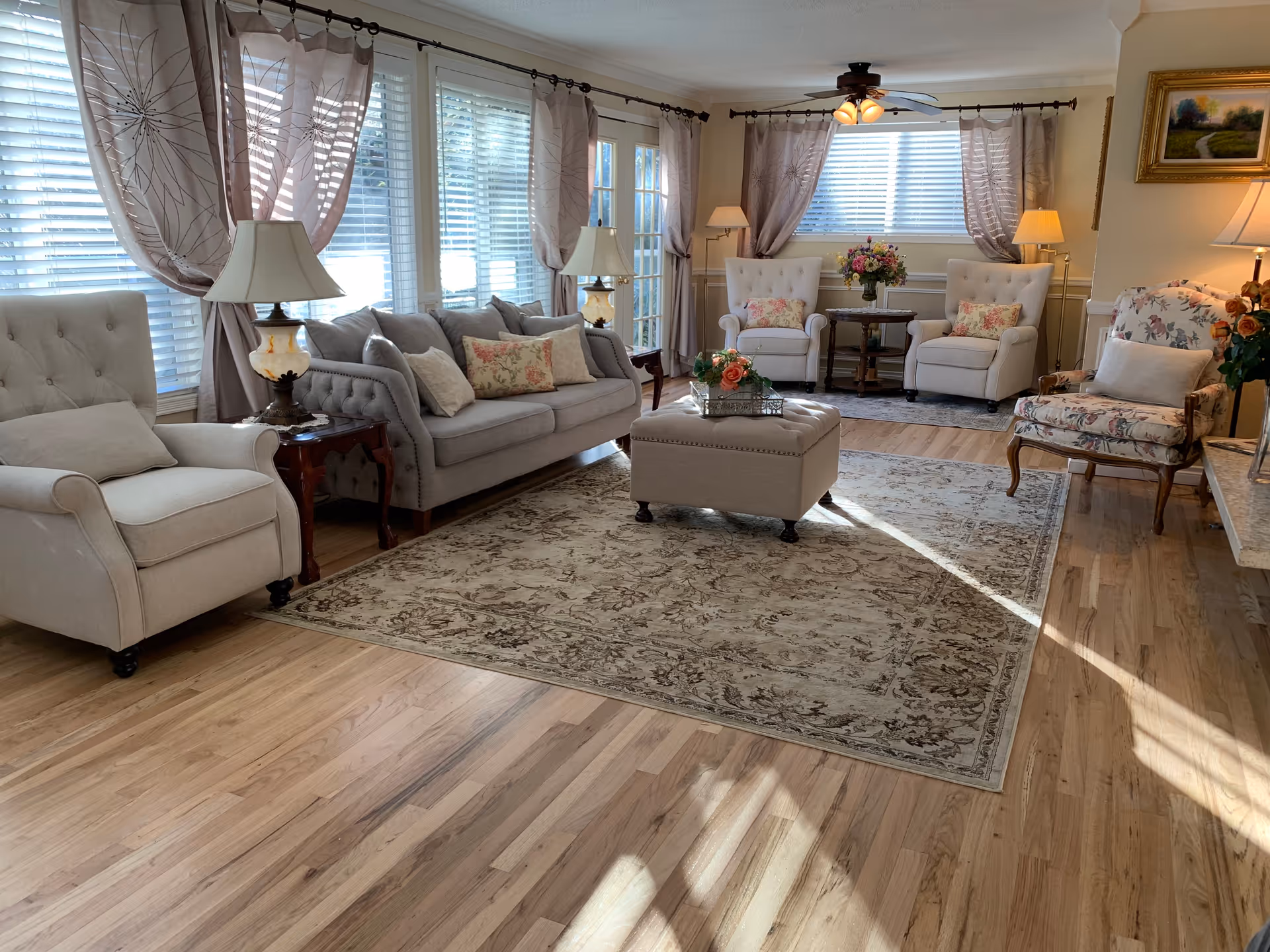 A bright and cozy living room with light wood flooring, a large patterned area rug, a beige sofa with floral and plain cushions, two beige armchairs, a floral-patterned armchair, and a beige ottoman in the center. The room has multiple windows with white blinds and sheer curtains, several table lamps, a ceiling fan with lights, and a small round table with a flower arrangement.
