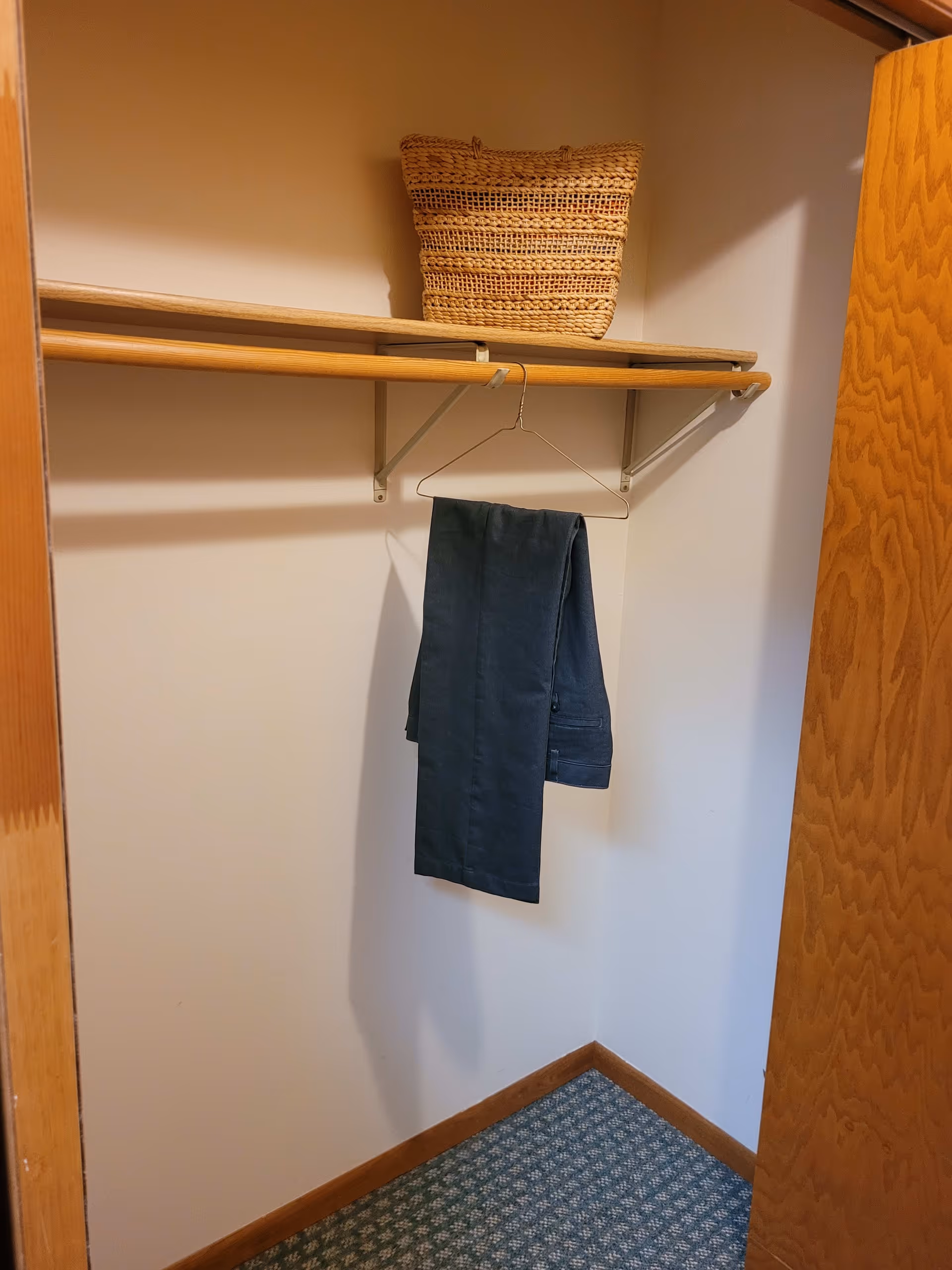 An empty closet with a wooden shelf and hanging rod. A woven basket is placed on the shelf, and a pair of dark pants is hanging on a wire hanger from the rod. The closet has beige walls, a wooden door partially open, and a patterned carpeted floor.
