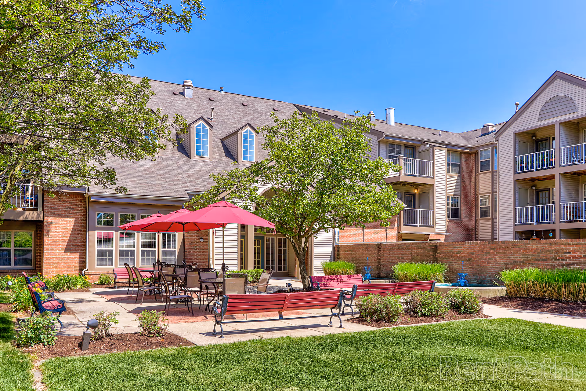 Outdoor courtyard area of Rosehaven Manor featuring red benches, tables with red umbrellas, a tree, and a multi-story building with balconies in the background under a clear blue sky.