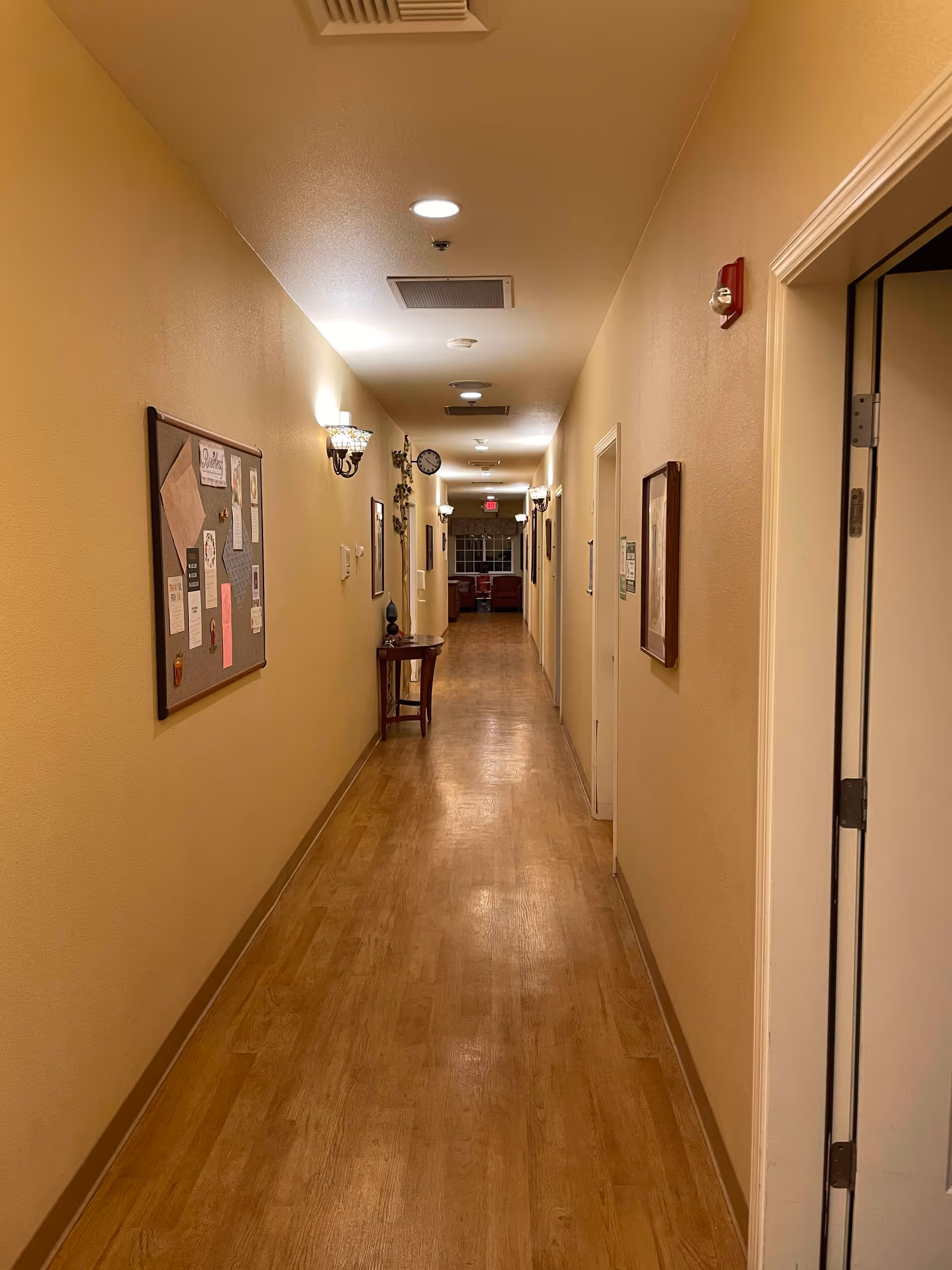 A long, well-lit hallway with wooden flooring and beige walls. The hallway has several doors on the right side and a bulletin board with various papers pinned on the left wall. Wall-mounted lights illuminate the corridor, and a small table with decorative items is placed along the left wall. At the end of the hallway, there is a window and some furniture visible.