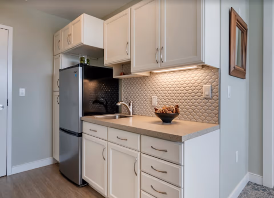 Small kitchenette with a stainless steel refrigerator, sink, light-colored cabinets, and a textured tile backsplash.