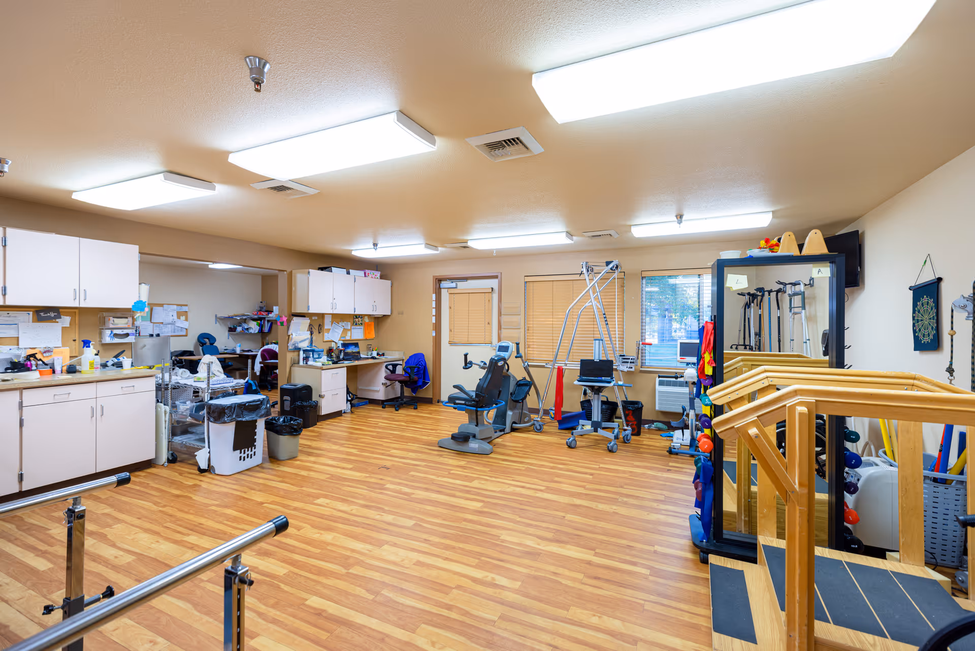 A spacious room with wooden flooring and beige walls, equipped with various physical therapy and exercise equipment including parallel bars, a stationary bike, and resistance bands. There are cabinets, desks with office chairs, and a large mirror on one side of the room. The room is well-lit with ceiling lights and has windows with blinds.