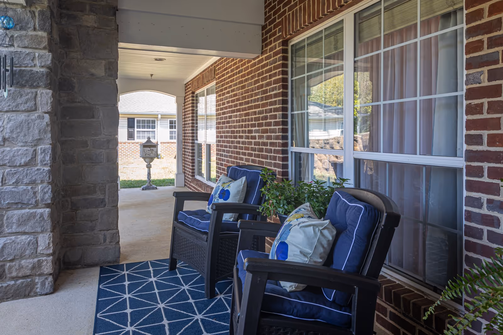 Covered outdoor patio area with two cushioned chairs featuring blue and white pillows, a blue geometric patterned rug, potted plants, and a brick wall with large windows.