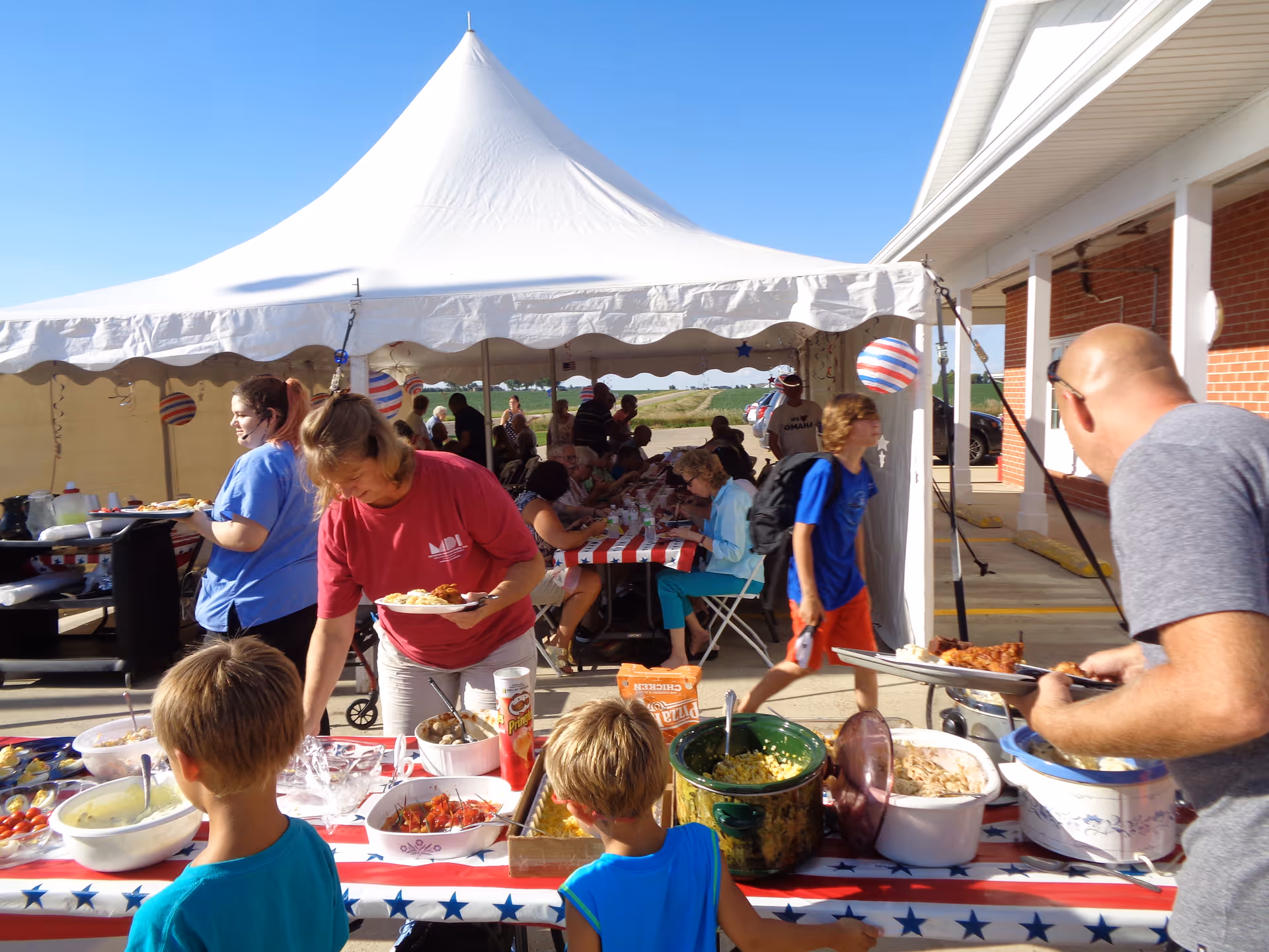 A group of people serve and eat food under a large white tent at an outdoor buffet table decorated with a red, white, and blue tablecloth.