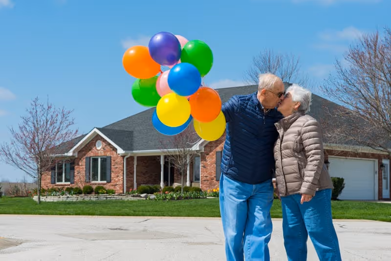 An elderly couple wearing jackets share a kiss outdoors in front of a brick house with a garage. The man holds a bunch of colorful balloons including orange, purple, green, blue, yellow, and pink. The sky is clear and blue with some trees in the background.