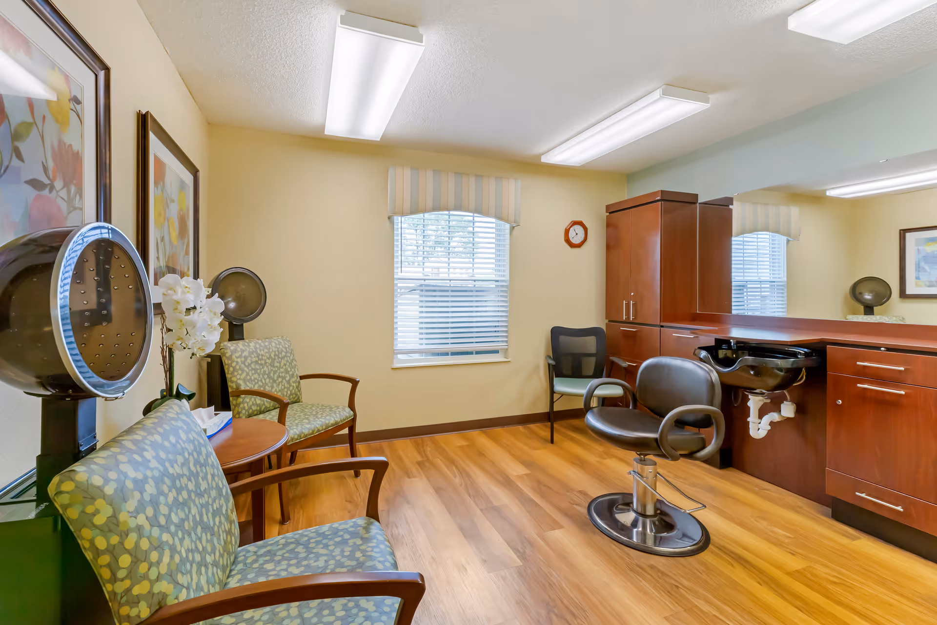 A well-lit hair salon area inside a senior living facility with wooden flooring, two green patterned chairs, a black salon chair in front of a counter with a built-in sink, a window with blinds and a striped valance, and framed floral artwork on the wall.