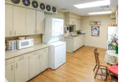 A bright kitchen area in a senior living facility featuring light wood cabinets, a microwave, a stacked washer and dryer, a sink, and a small dining table with chairs. The floor is wood, and there are decorative plates displayed above the cabinets. A window lets in natural light.