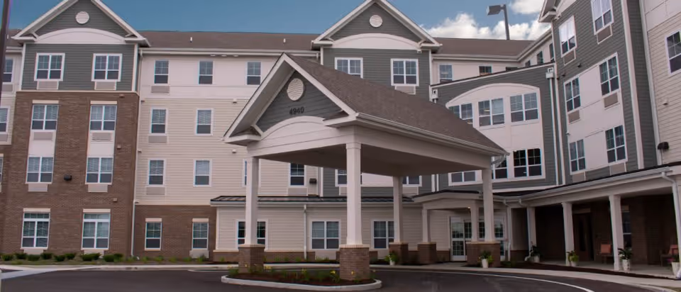 Front exterior view of a multi-story senior living facility named Oasis at 56th, featuring a covered entrance with pillars and a circular driveway, surrounded by windows and a mix of brick and siding on the building facade under a partly cloudy sky.