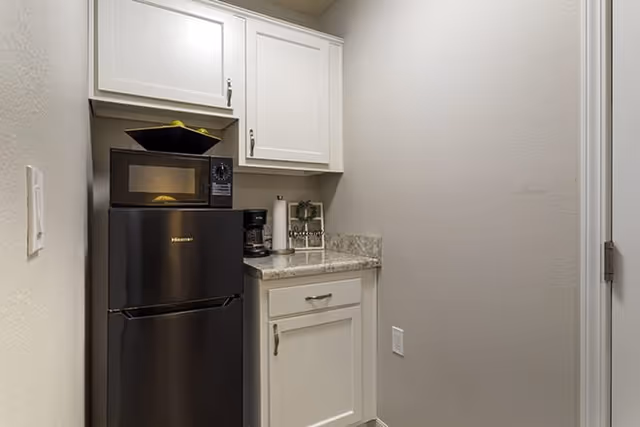 Small kitchenette area with a black refrigerator, microwave on top, white cabinets above and below a granite countertop, a coffee maker, paper towel holder, and a small decorative plant on the counter.