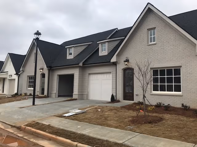 Front exterior of attached cottage-style residences with garages, a lamppost, and newly landscaped lawns under an overcast sky.