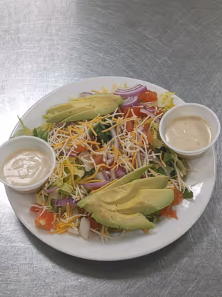 Plate of salad with avocado slices, shredded cheese, red onion, tomatoes, and two cups of dressing on a stainless steel surface.