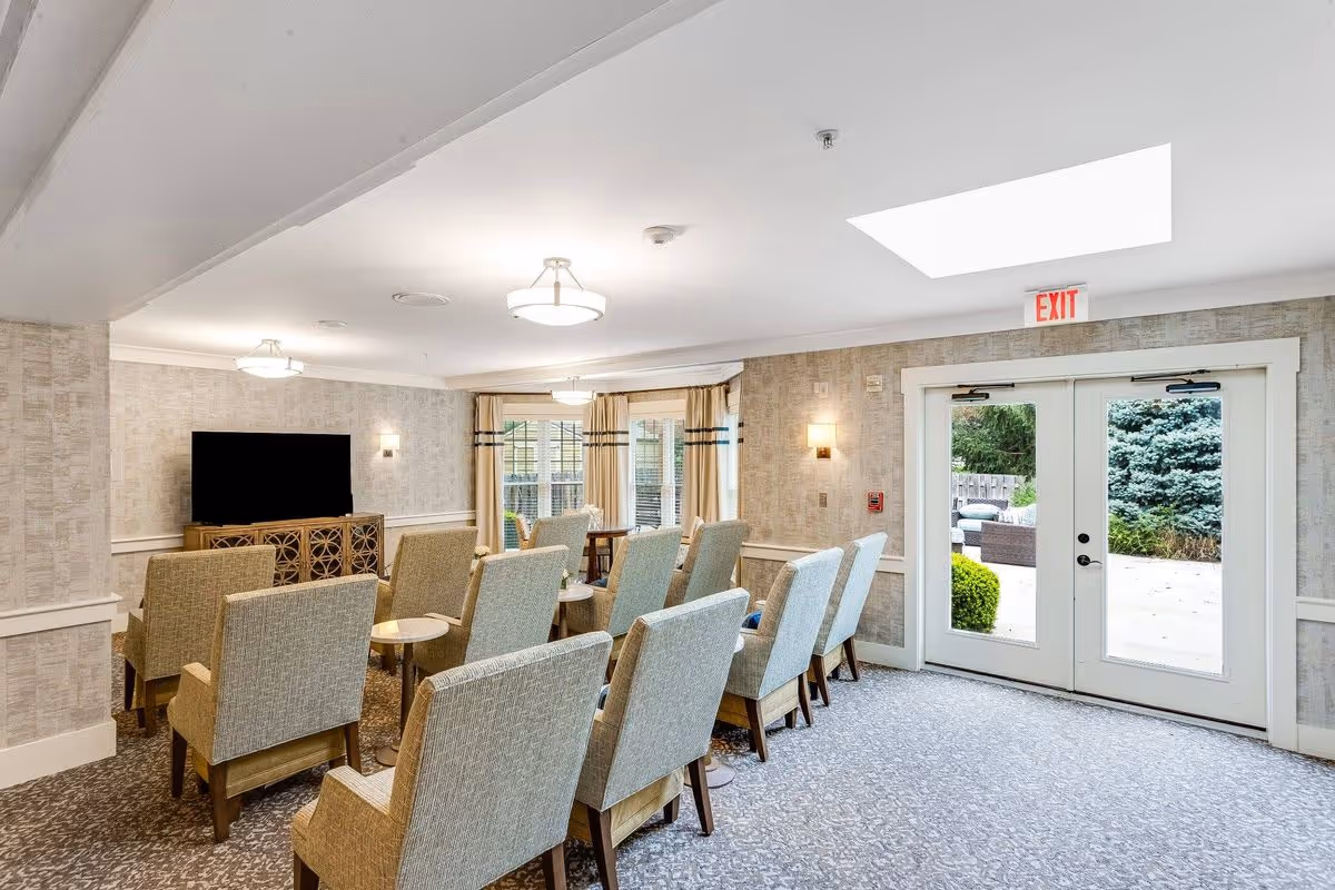 A bright and spacious common room with multiple beige upholstered chairs arranged in rows facing a large flat-screen TV on a wooden cabinet. The room has patterned carpet flooring, light-colored wallpaper, ceiling lights, and large windows with curtains letting in natural light. Double glass doors lead outside to a patio area with greenery visible.