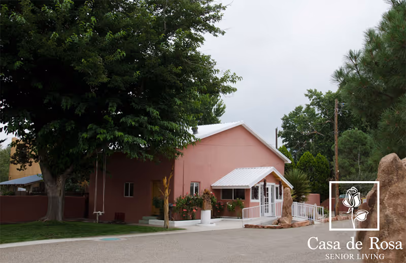 Exterior view of Casa de Rosa Assisted Living facility showing a pink building with a white roof, surrounded by trees and greenery, with a paved driveway in front.